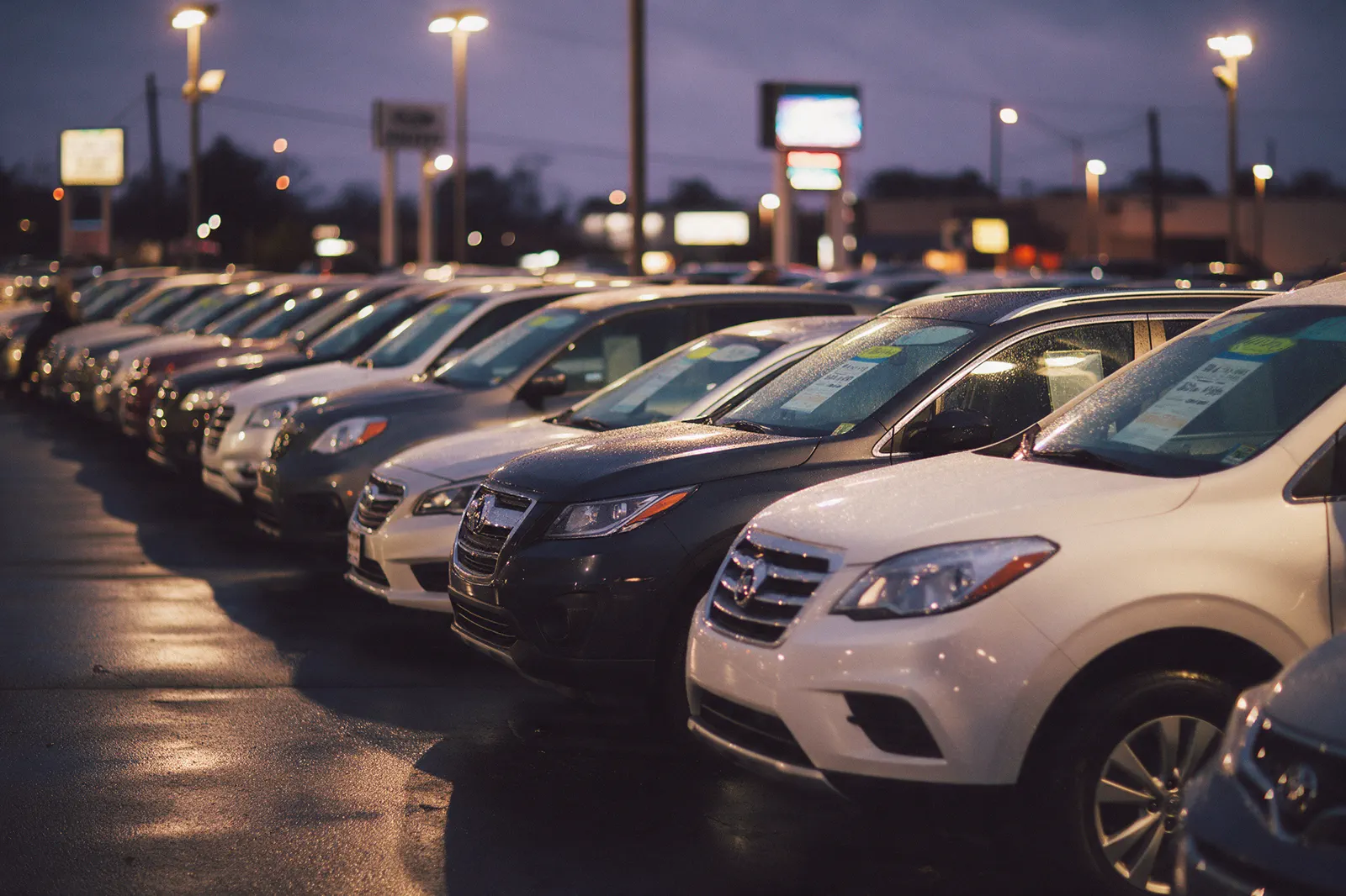 Dealership front lot at dusk, a row of vehicles with price tags going soft toward the back.