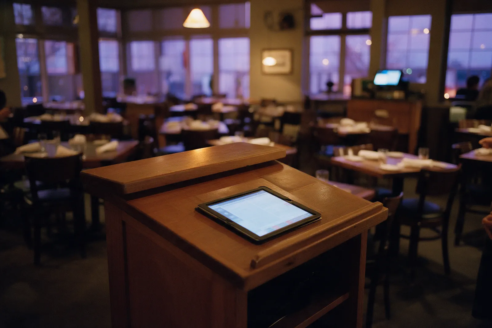 Restaurant hostess stand at dusk, the reservation tablet lit over an empty dining room.